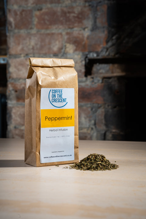 A package of peppermint tea leaves next to a small pile of dried leaves, with a label on the package that reads 'Peppermint'. The package is sealed and has Coffee On The Cresent branding visible.